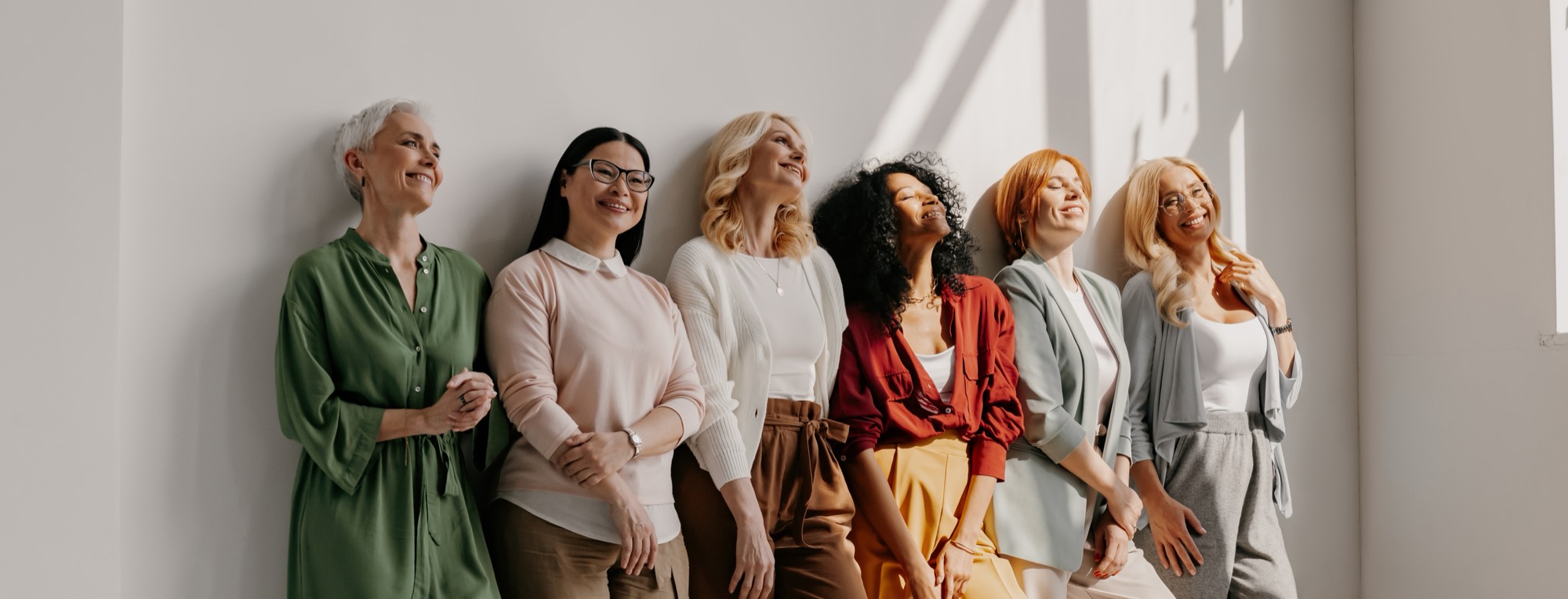 Six women of different generations standing together against a sunlit wall