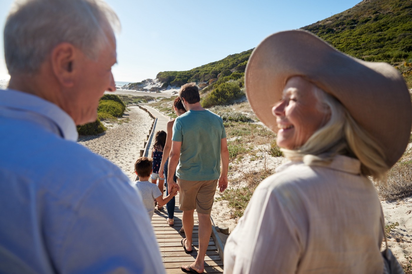 Three generations of a family walking along a wooden boardwalk through coastal dunes toward the sea, with a smiling older man and woman in the foreground