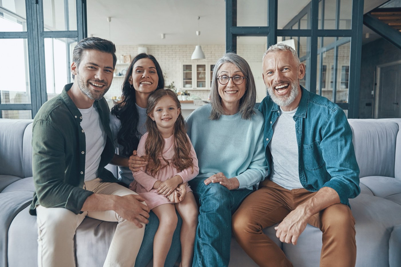 A smiling multigenerational family of four adults and a young girl sitting close together on a sofa in a bright living room