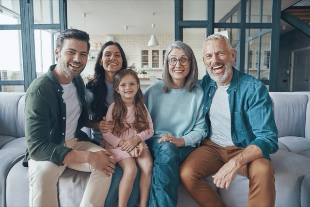 A smiling multigenerational family of four adults and a young girl sitting close together on a sofa in a bright living room