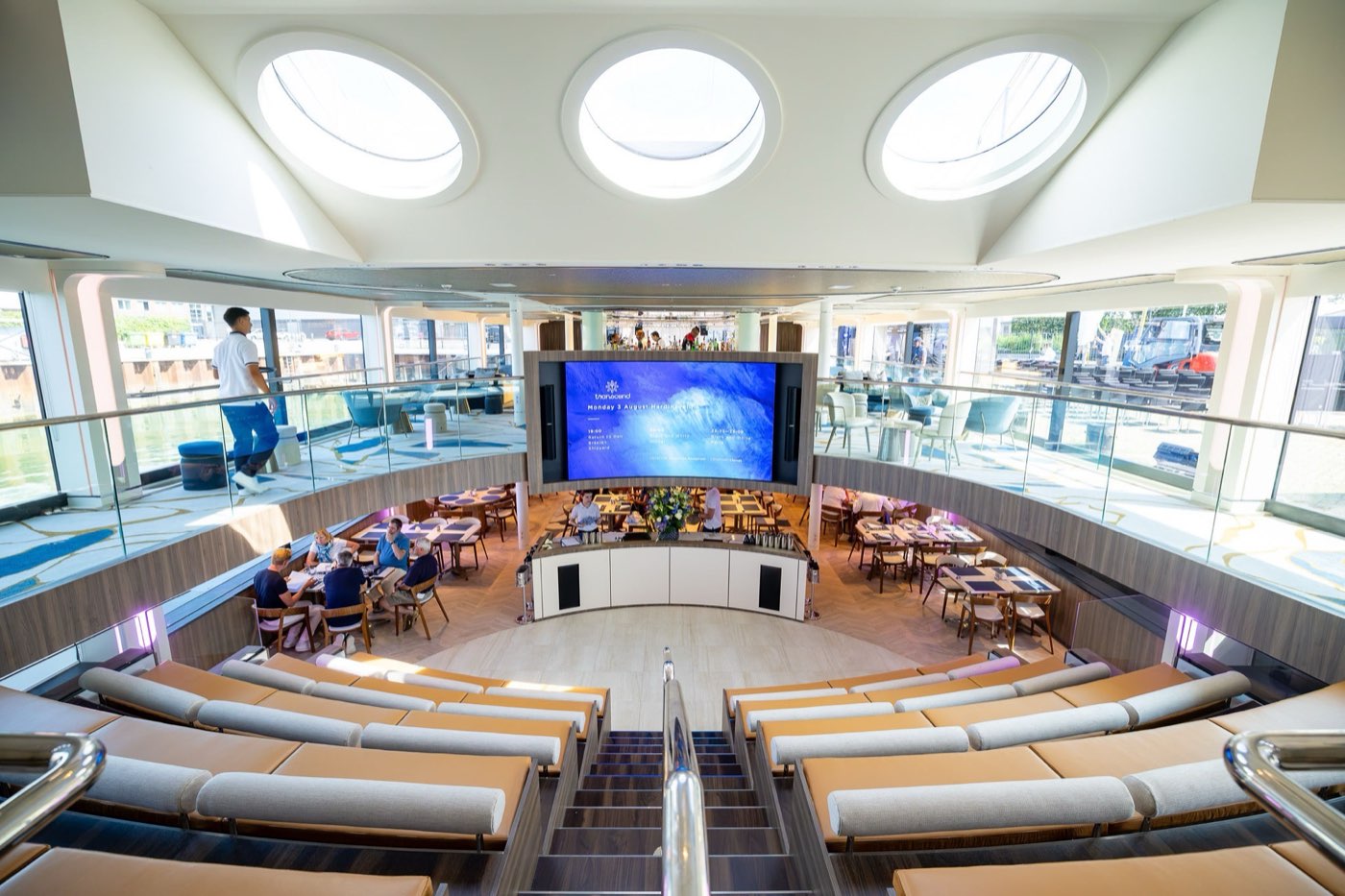 Tiered lounge seating facing a large screen in a ship's atrium, with round skylights in the ceiling
